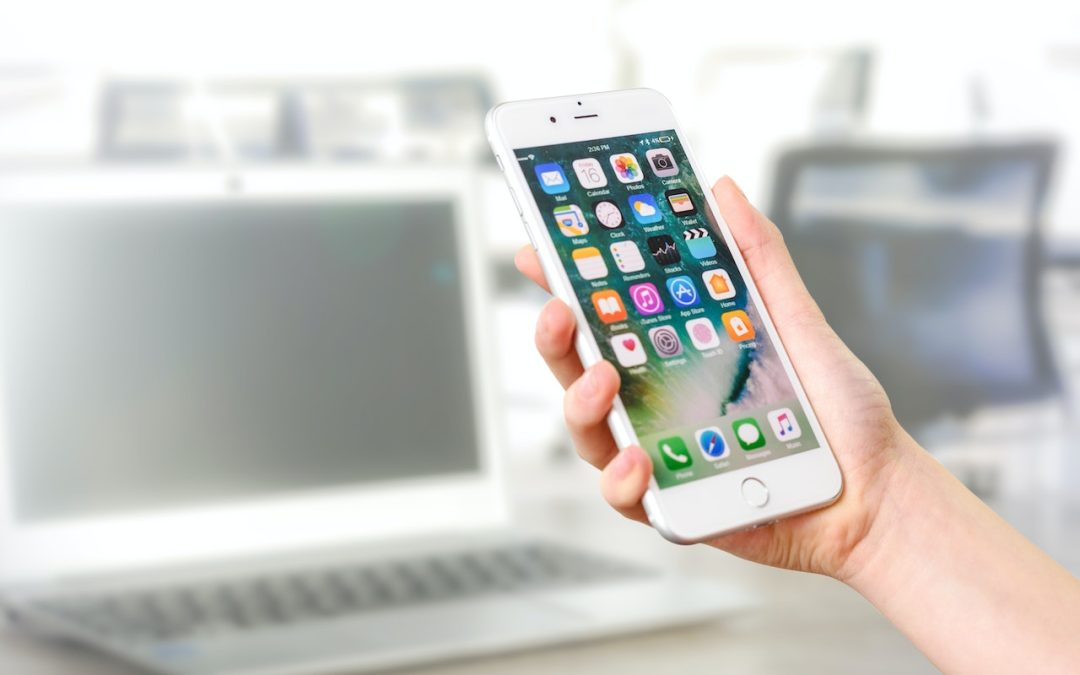 Feature photo description. This is a picture of a right hand holding a white cell phone. It also has a blurred background featuring an open laptop